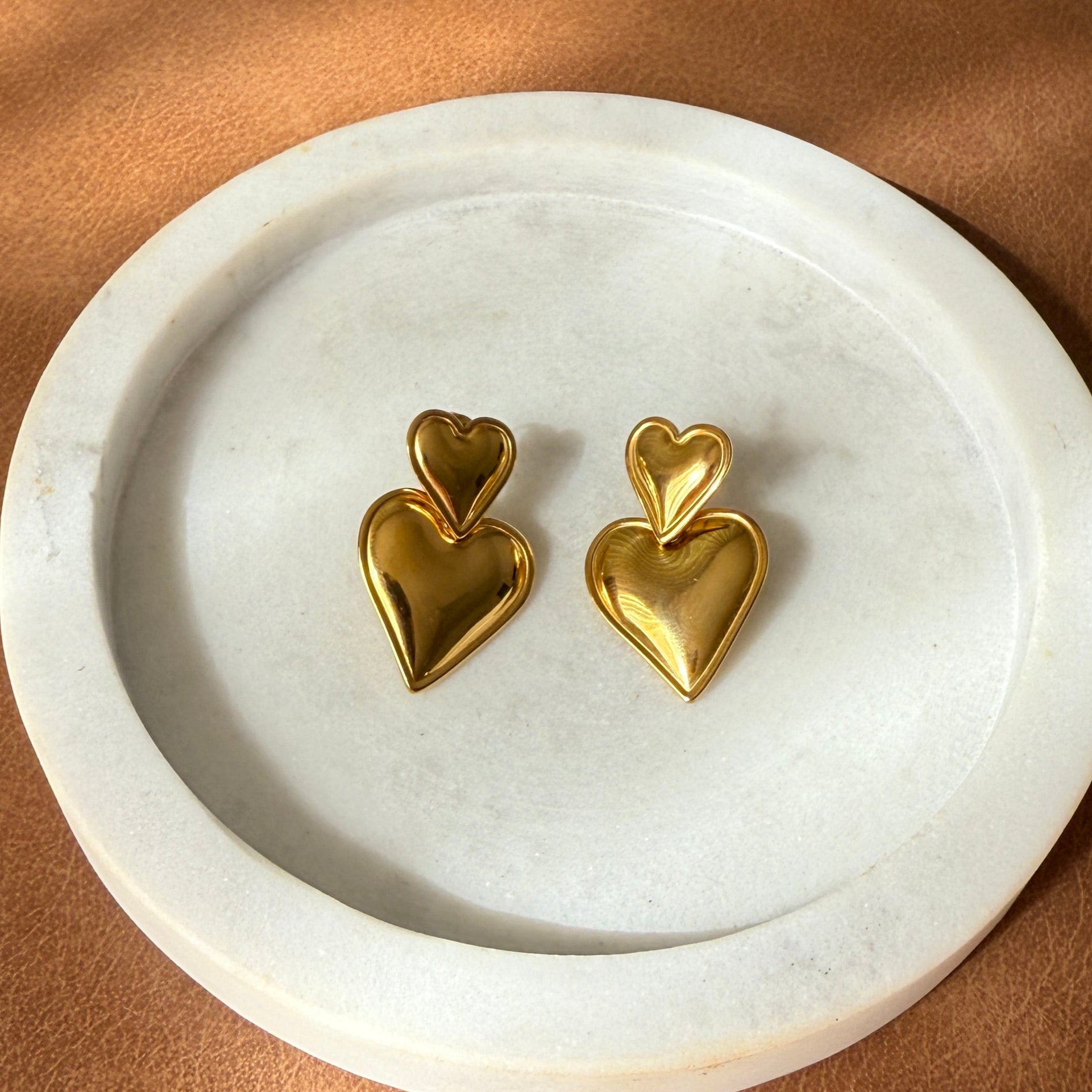 Gold heart-shaped earrings on a white circular stand against a brown leather background
